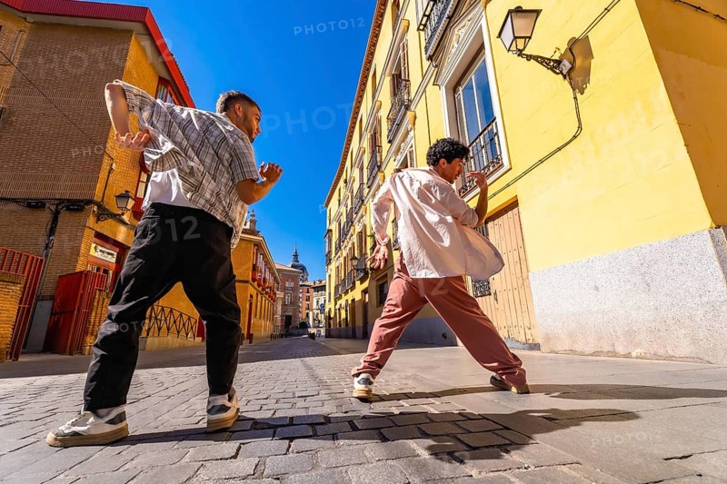 Two young men expressing themselves through street dance movement, performing a modern hip hop choreography on a cobblestone road in an old european city street under bright sunlight