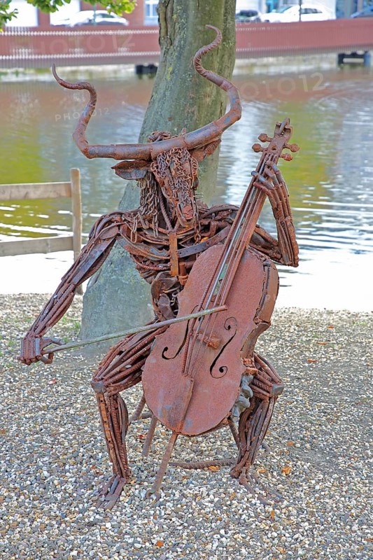 Sculpture The Concert by Günther Fahrner, rusty metal sculpture, sheet metal, modern art, bizarre whimsical musician sitting in a chair, cello player, hybrid creature, playing music, pond, beach, shore, bull figure, Bastinsweiher, Rhineland, Stolberg, North Rhine-Westphalia, Germany