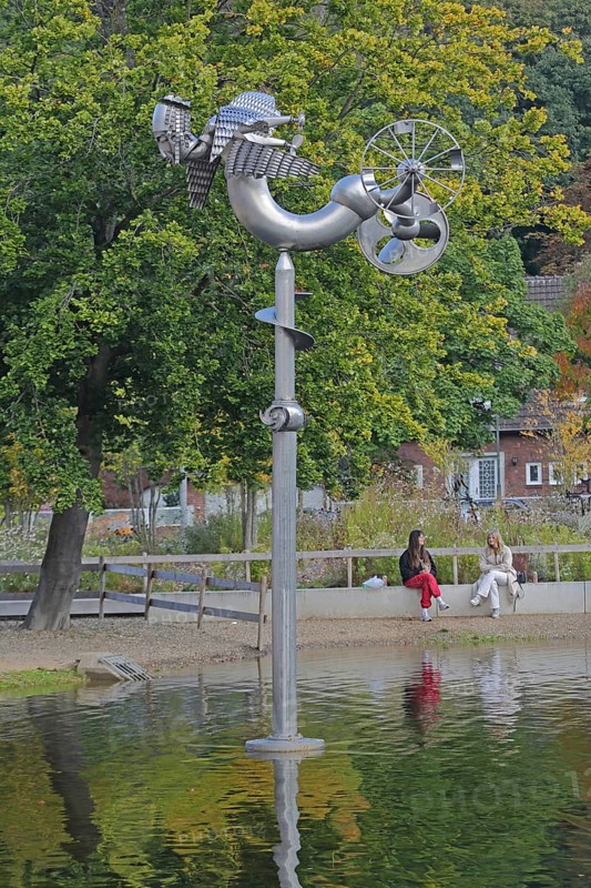 Sculpture Birdman by Albert Sous and two seated woman in conversation, people, pond, modern art, abstract art, stainless steel, metal sculpture, bizarre bizarre bird figure, mythical creature, hybrid creature, wings, flying, wheel, free-standing, Bastinsweiher, Rhineland, Stolberg, North Rhine-Westphalia, Germany