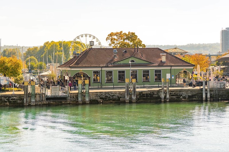 A building at the harbour with autumnal vegetation and a Ferris wheel in the background in a cityscape, Constance, Lake Constance, Germany