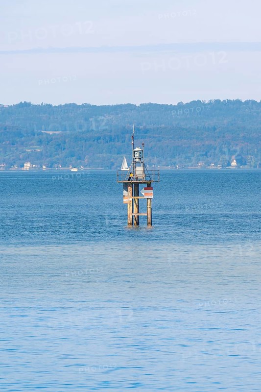 A lighthouse in blue water with a sailing ship, green hills and clear sky in the background, Constance, Lake Constance, Germany