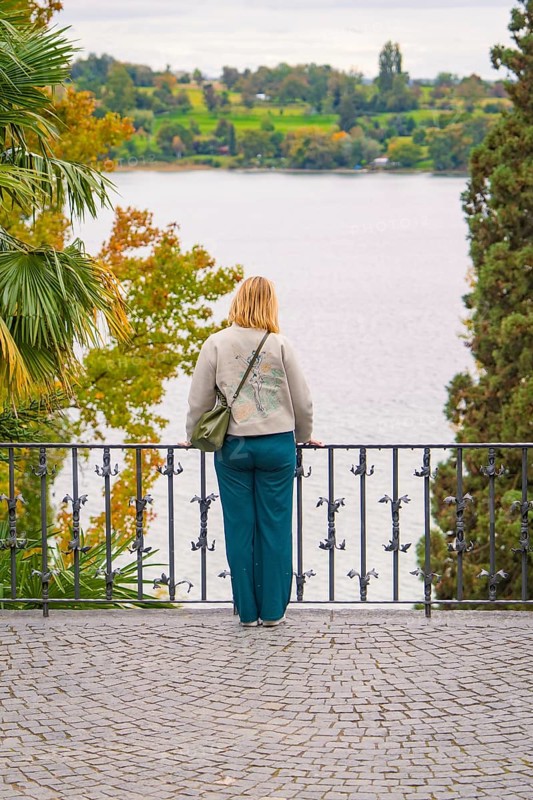 Person standing at the railing of a walkway and looking at a lake surrounded by trees in autumn, Mainau Island, Lake Constance, Germany