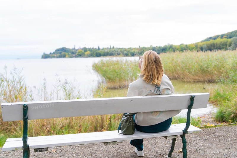 Woman on park bench looking at calm lake and autumn landscape, Mainau Island, Lake Constance, Germany