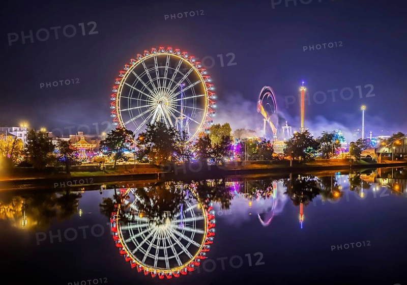 Folk festival in Stuttgart. Night shot with light effects. The 178th Cannstatter Volksfest on the Wasen is one of the most important traditional festivals in Germany. The Neckar River reflects the Ferris wheel and other attractions of the amusement park. Stuttgart, Baden-Württemberg, Germany