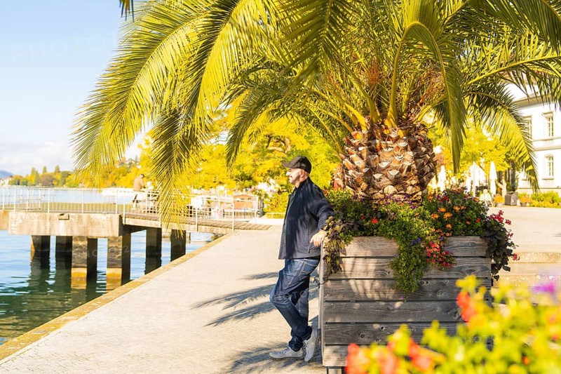 A man leaning against a flower trough on a sunny promenade by the water, Überlingen, Lake Constance, Germany