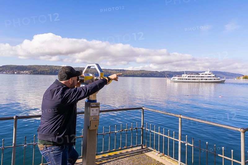 Man pointing with binoculars at a ship on a calm lake under a clear sky, Überlingen, Lake Constance, Germany
