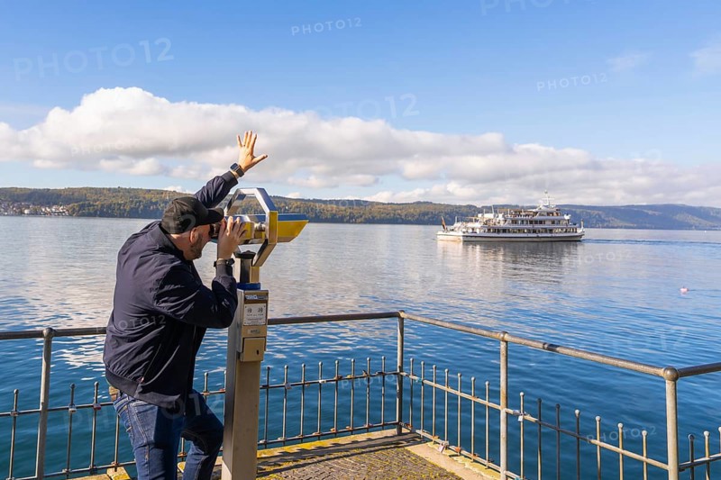 A man uses a telescope to observe a ship on a calm lake under a clear sky, Überlingen, Lake Constance, Germany