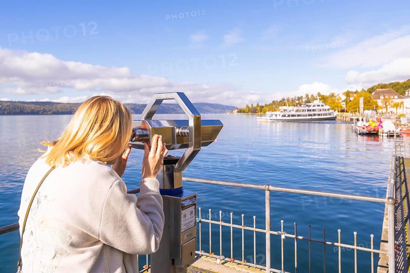 A woman looks through a telescope at a lake with ships and a clear autumn sky, Überlingen, Lake Constance, Germany