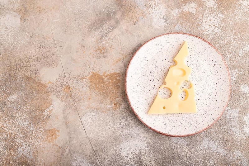 Piece of cheese with holes on ceramic plate on brown concrete background, top view, flat lay, copy space, minimalism