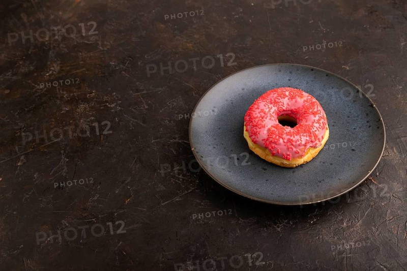 Pink Donut with sprinkles on blue ceramic plate on black concrete background, side view, copy space, minimalism