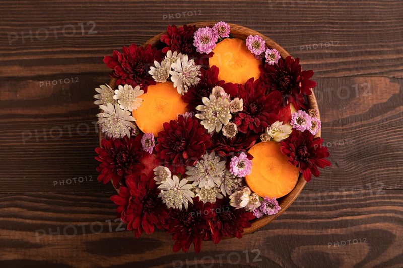 Wooden bowl with carrot slices and red Chrysanthemum flowers, Astrantia flowers, flower salad on brown wooden background, top view, flat lay, close up, minimalism