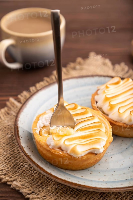 French lemon tart with meringue on brown wooden background, cup of coffee, linen textile, side view, close up, selective focus