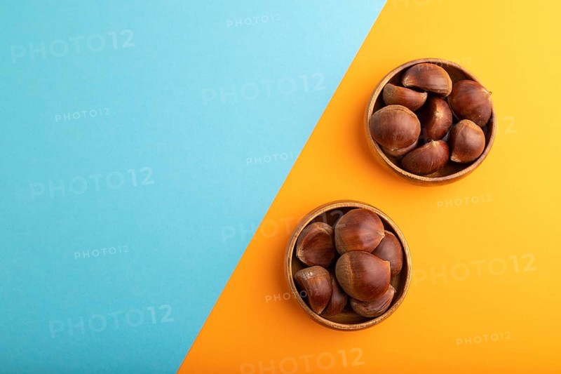 Wooden bowl with raw edible ?hestnuts on blue and orange paper pastel background, top view, flat lay, copy space, minimalism