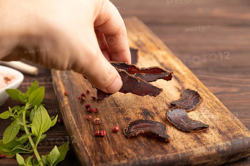Armenian Basturma dried meat with hand on wooden cutting board with pepper and herbs on brown wooden background. Side view, close up, selective focus