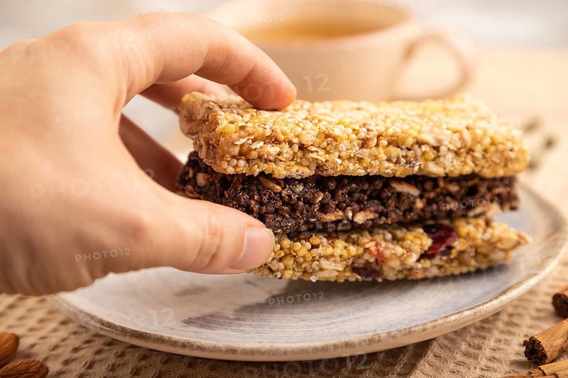 Granola bars with caramel, nuts, flakes in ceramic plate on brown concrete background with hand, beige linen napkin, cup of green tea. Side view, close up, selective focus