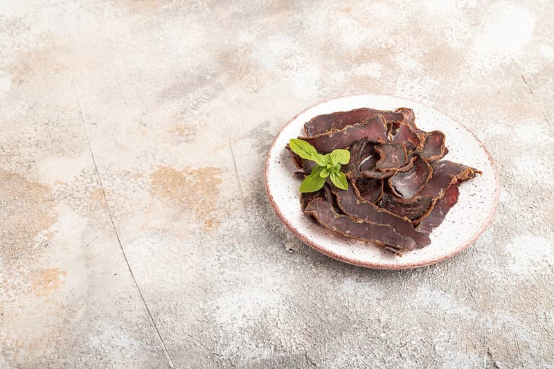 Armenian Basturma dried meat on plate with pepper and herbs on brown concrete background. Side view, copy space