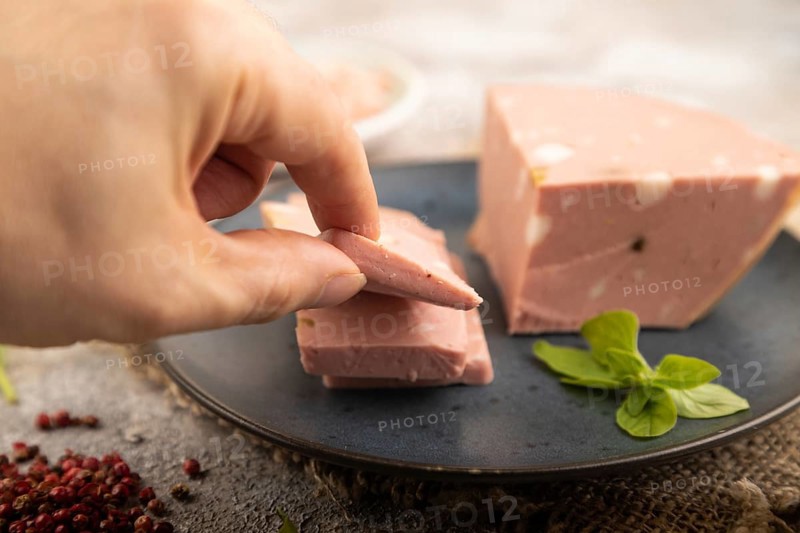 Mortadella sausage with hand on plate with pepper and herbs on brown concrete background and linen textile. Side view, close up, selective focus