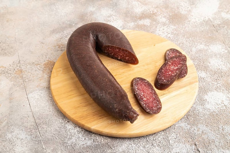 Sujuk sausage on wooden cutting board with pepper and herbs on brown concrete background. Side view, close up