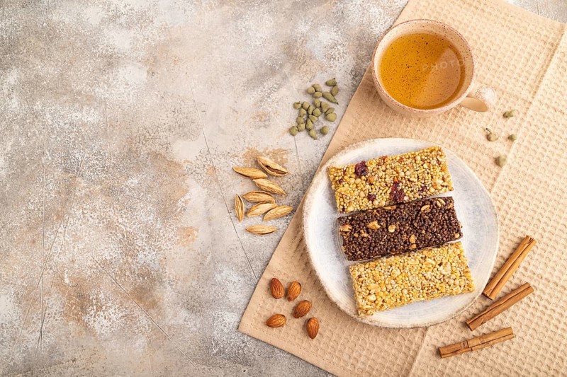 Granola bars with caramel, nuts, flakes in ceramic plate on brown concrete background, beige linen napkin, cup of green tea. Top view, flat lay, copy space