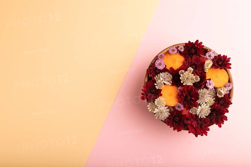Wooden bowl with carrot slices and red Chrysanthemum flowers, Astrantia flowers, flower salad on pink and orange pastel paper background, top view, flat lay, copy space, minimalism