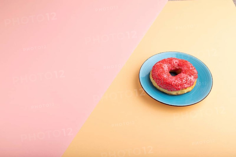 Pink Donut with sprinkles on blue ceramic plate on blue and orange pastel paper background, side view, copy space, minimalism