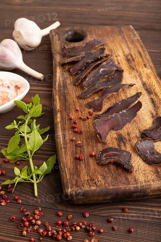Armenian Basturma dried meat on wooden cutting board with pepper and herbs on brown wooden background. Side view, close up