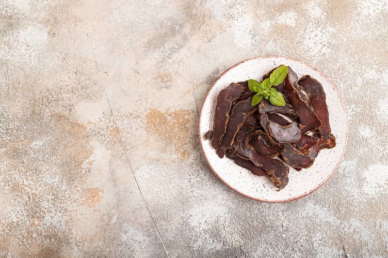Armenian Basturma dried meat on plate with pepper and herbs on brown concrete background. Top view, flat lay, copy space