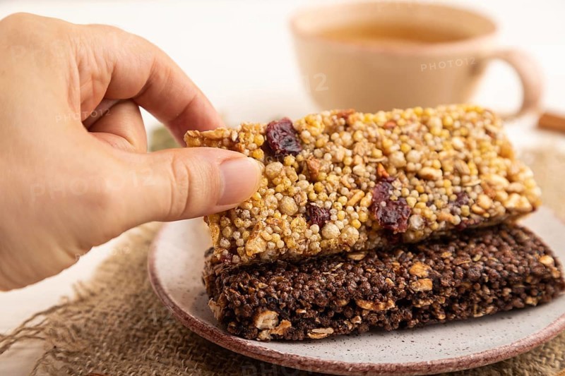 Granola bars with caramel, nuts, flakes in ceramic plate with hand on white wooden background, beige linen napkin, cup of green tea. Side view, close up, selective focus