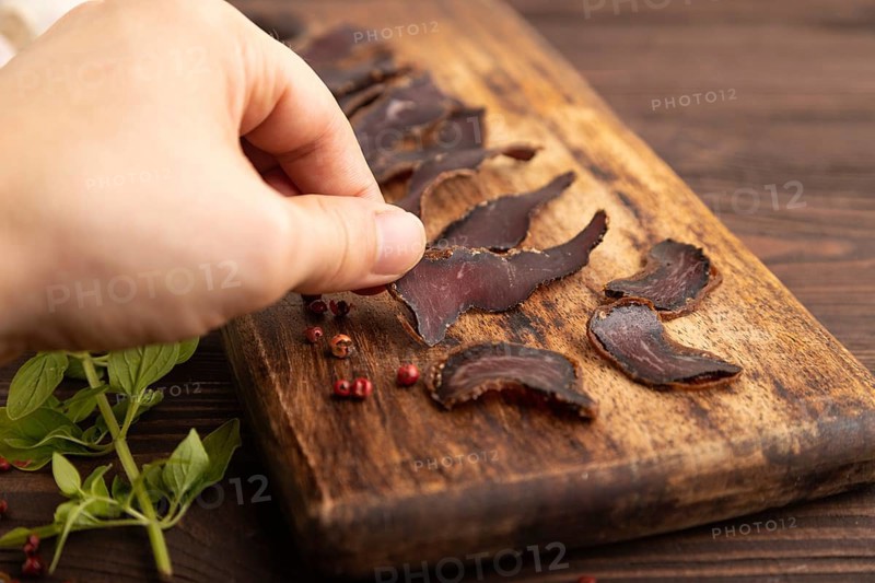 Armenian Basturma dried meat with hand on wooden cutting board with pepper and herbs on brown wooden background. Side view, close up, selective focus