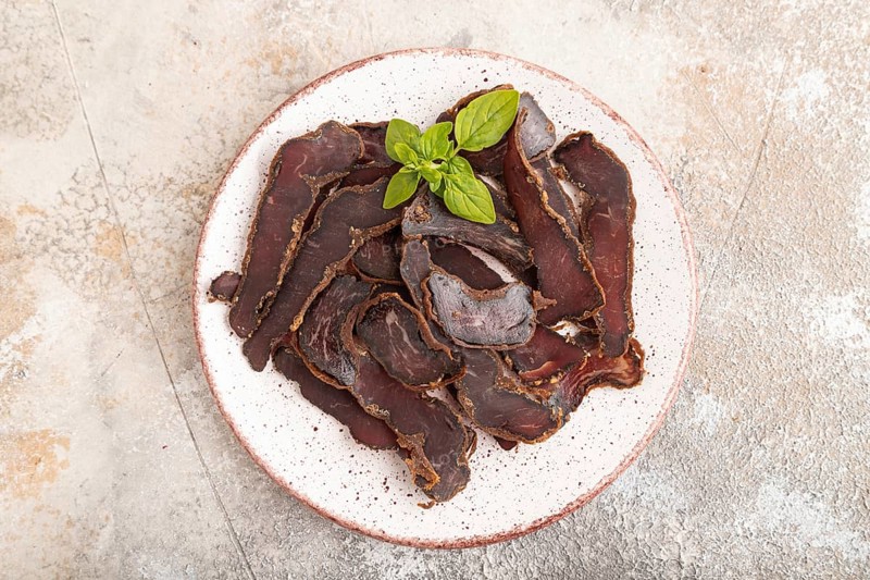 Armenian Basturma dried meat on plate with pepper and herbs on brown concrete background. Top view, flat lay, copy space