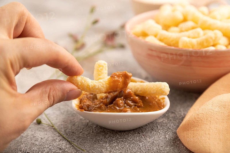 Corn flakes sticks with hand with caramel in ceramic bowl on gray concrete background and orange linen textile. Side view, close up