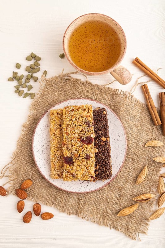 Granola bars with caramel, nuts, flakes in ceramic plate on white wooden background, beige linen napkin, cup of green tea. Top view, flat lay, close up