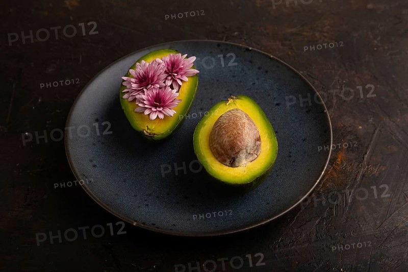 Two Avocado halves with pit and Chrysanthemums on blue plate on black concrete background, side view, minimalism, low key, close up