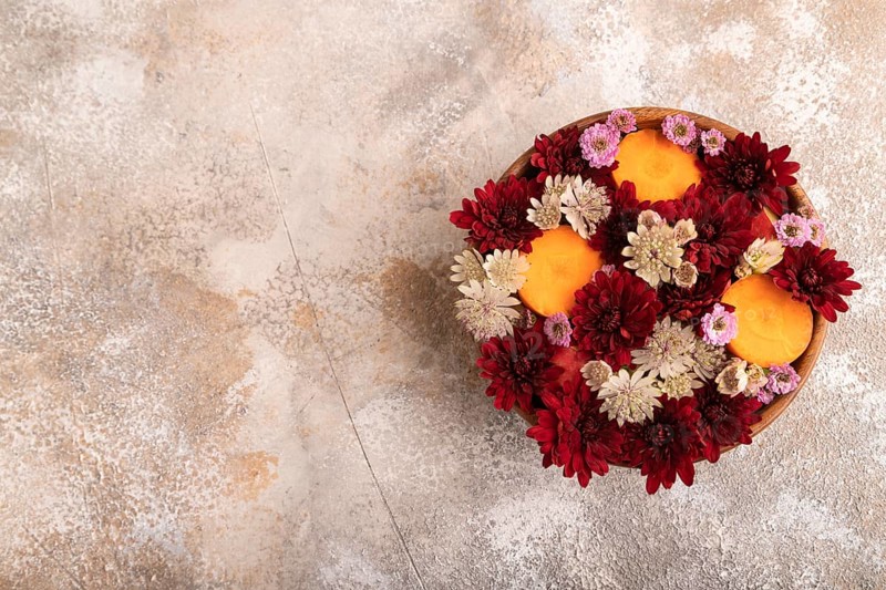 Wooden bowl with carrot slices and red Chrysanthemum flowers, Astrantia flowers, flower salad on brown concrete background, top view, flat lay, copy space, minimalism