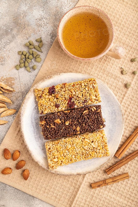 Granola bars with caramel, nuts, flakes in ceramic plate on brown concrete background, beige linen napkin, cup of green tea. Top view, flat lay, close up