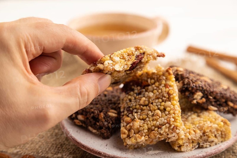 Granola bars with caramel, nuts, flakes in ceramic plate with hand on white wooden background, beige linen napkin, cup of green tea. Side view, close up, selective focus