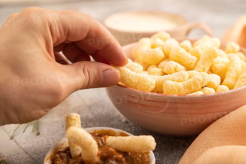 Corn flakes sticks with hand with caramel in ceramic bowl on gray concrete background and orange linen textile. Side view, close up