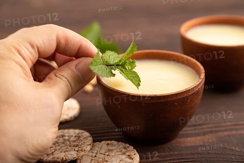 Organic non dairy banana and soy milk in clay cup with hand on brown wooden background. Vegan healthy food concept, side view, close up, selective focus