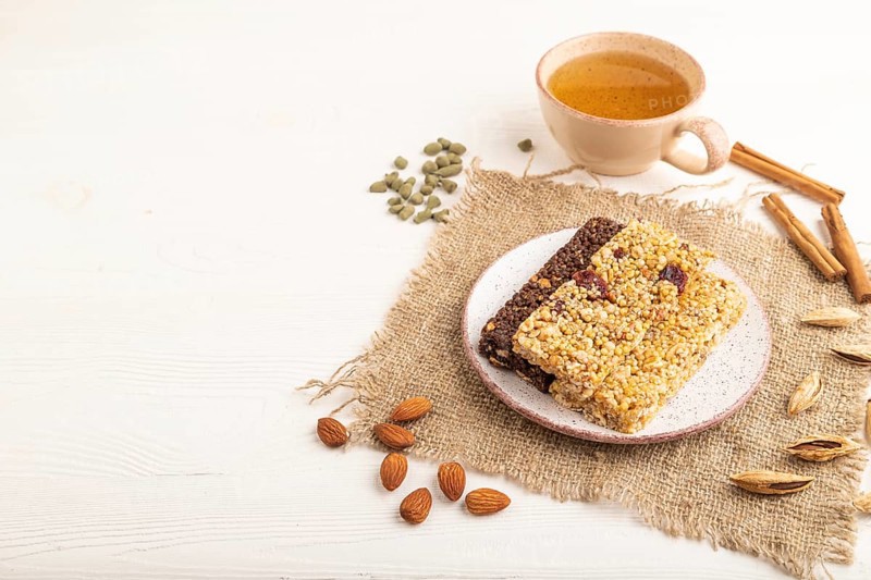 Granola bars with caramel, nuts, flakes in ceramic plate on white wooden background, beige linen napkin, cup of green tea. Side view, copy space