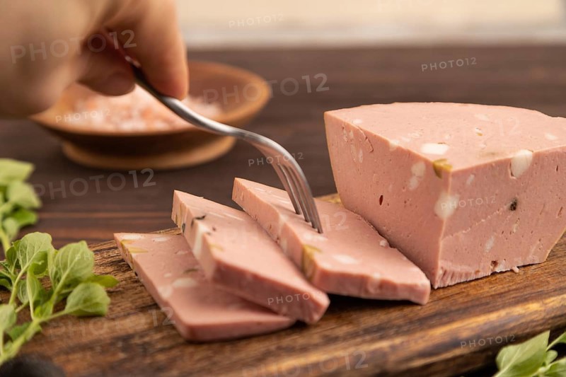 Mortadella sausage with hand on wooden cutting board with pepper and herbs on brown wooden background. Side view, close up, selective focus