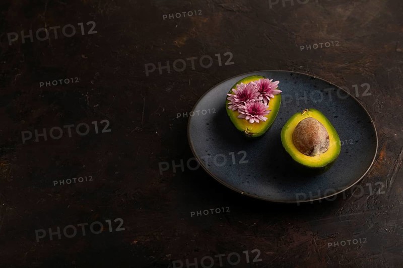 Two Avocado halves with pit and Chrysanthemums on blue plate on black concrete background, side view, copy space, minimalism, low key