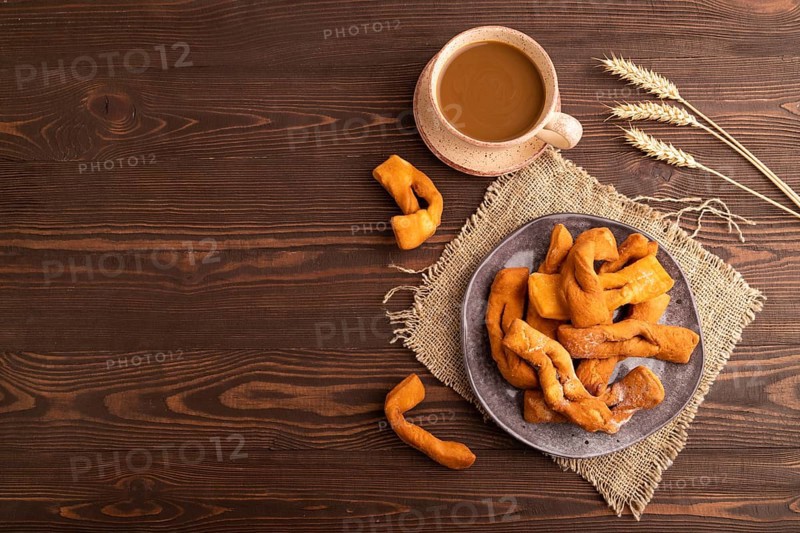 Crunchy biscuit Brushwood cookies sprinkled with powdered sugar on brown wooden background and linen textile, cup of coffee, top view, flat lay, copy space