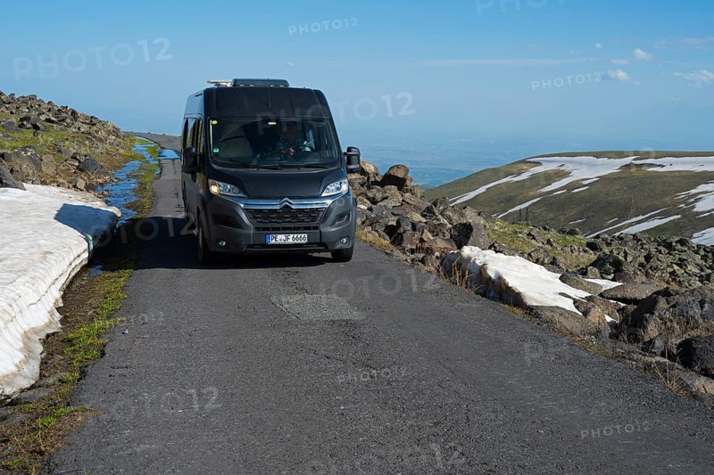 Van fährt auf asphaltiertem Weg neben Felsen und Schneefeldern mit klarem Himmel, das Tal im Hintergrund, mit dem Camper zum Aragaz, Aragats, Aragac, Alagyaz, erloschener Schichtvulkan, 4090 m, Aragazotn, Schirak, Armenien