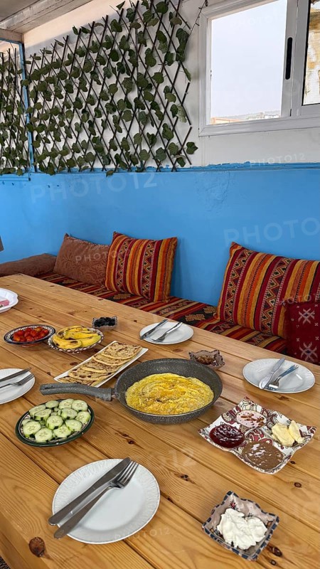 Rustic wooden table features a spread of moroccan breakfast food, with a pan of omelette, flatbread, fresh vegetables, various spreads, and vibrant textiles on the seating area