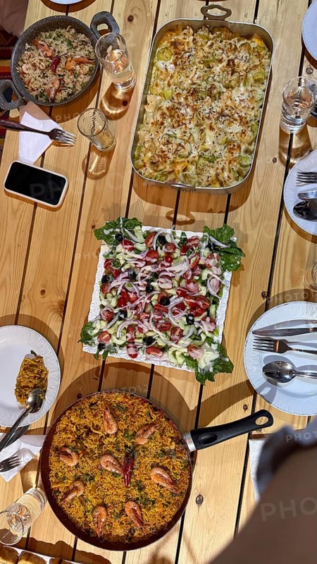 Rustic wooden table set with multiple homemade dishes including a large pan of seafood paella, a casserole, and a fresh salad, creating a vibrant feast for sharing during a meal