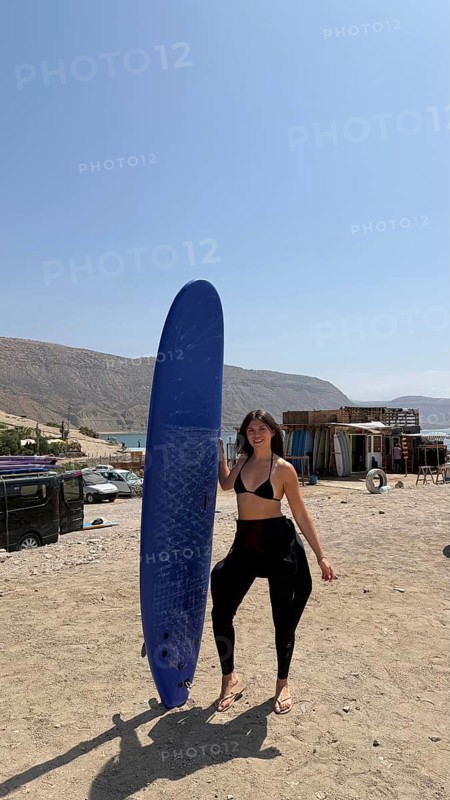 Young woman on a sunny moroccan beach holding a blue surfboard, wearing a bikini top and wetsuit bottoms, smiling and preparing to surf in calm ocean waters near taghazout
