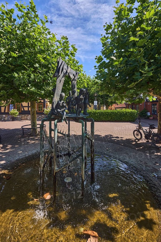 Brunnen mit Metallskulpturen, Alltägliches Theater, Bildhauer Hugo Uhl, Platz, Bäume, Hecke, blauer Himmel, Cirrostratuswolken, Burgstraße, Hofheim am Taunus, Main-Taunus-Kreis, Hessen, Deutschland