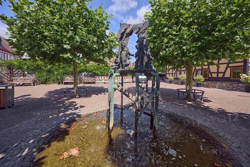 Brunnen mit Metallskulpturen, Alltägliches Theater, Bildhauer Hugo Uhl, Platz, Bäume, Hecke, schattig, blauer Himmel, Cirrostratuswolken, Kreuzung zwischen Tivertonplatz, Burgstraße, Burggrabenstraße und Taubengasse, Hofheim am Taunus, Main-Taunus-Kreis, Hessen, Deutschland