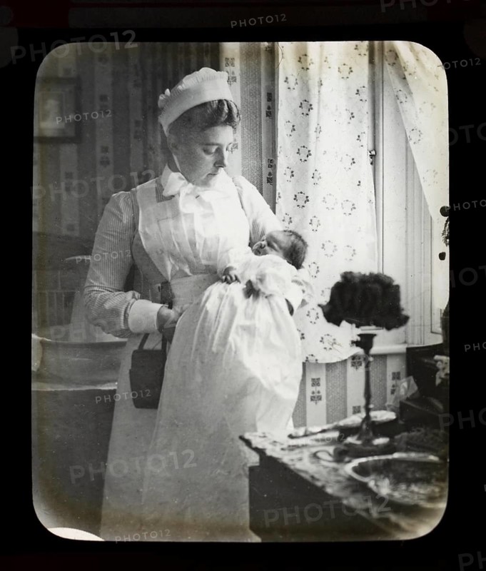 Magic lantern slide of nurse in uniform holding newborn baby inside a home, England, UK c 1900-1910