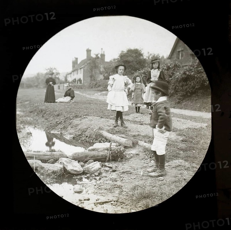 Magic lantern slide of mothers with children outside playing by a stream in a village rural location, UK circa 1900